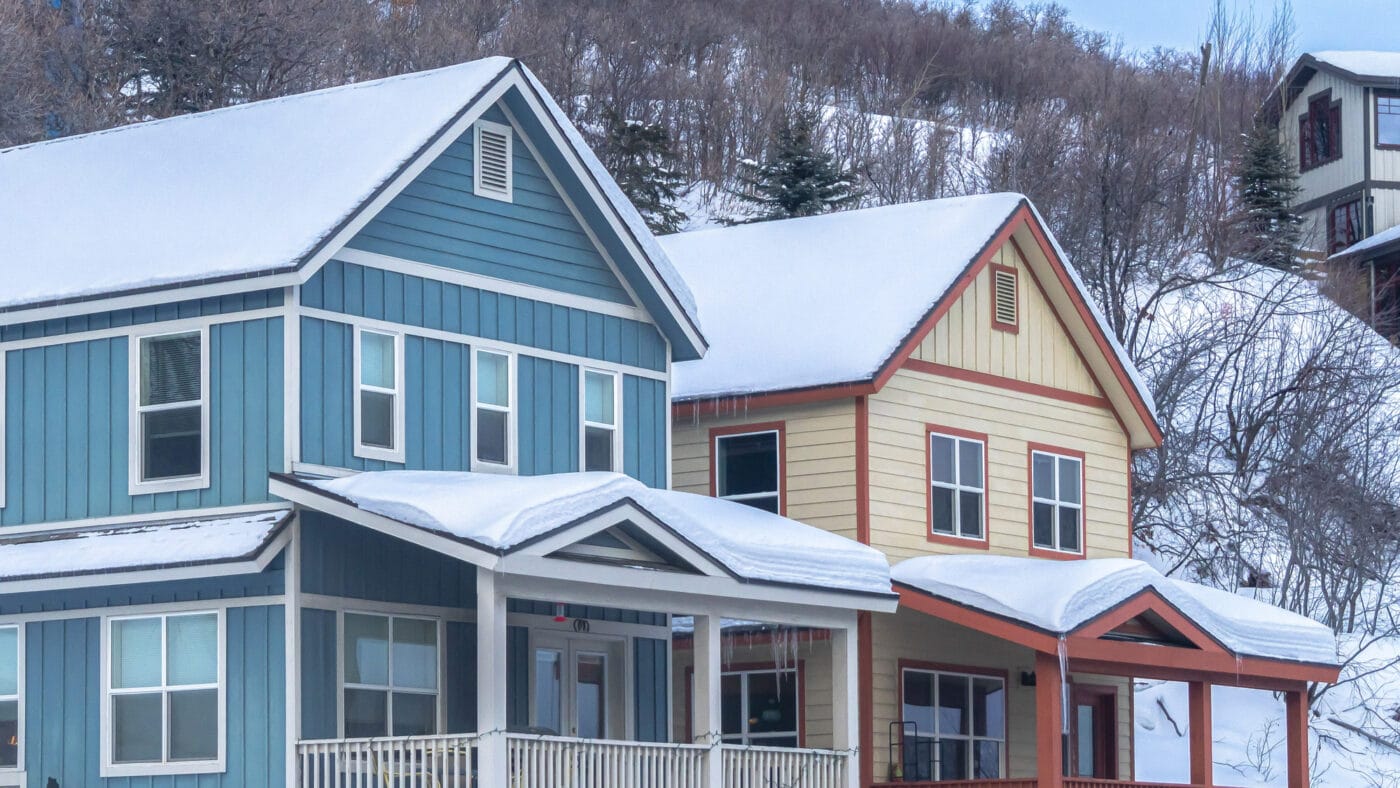Two snow-covered houses in a wintry landscape, one blue with white trim and a porch, the other cream with red trim, both surrounded by snow-covered trees and slopes.