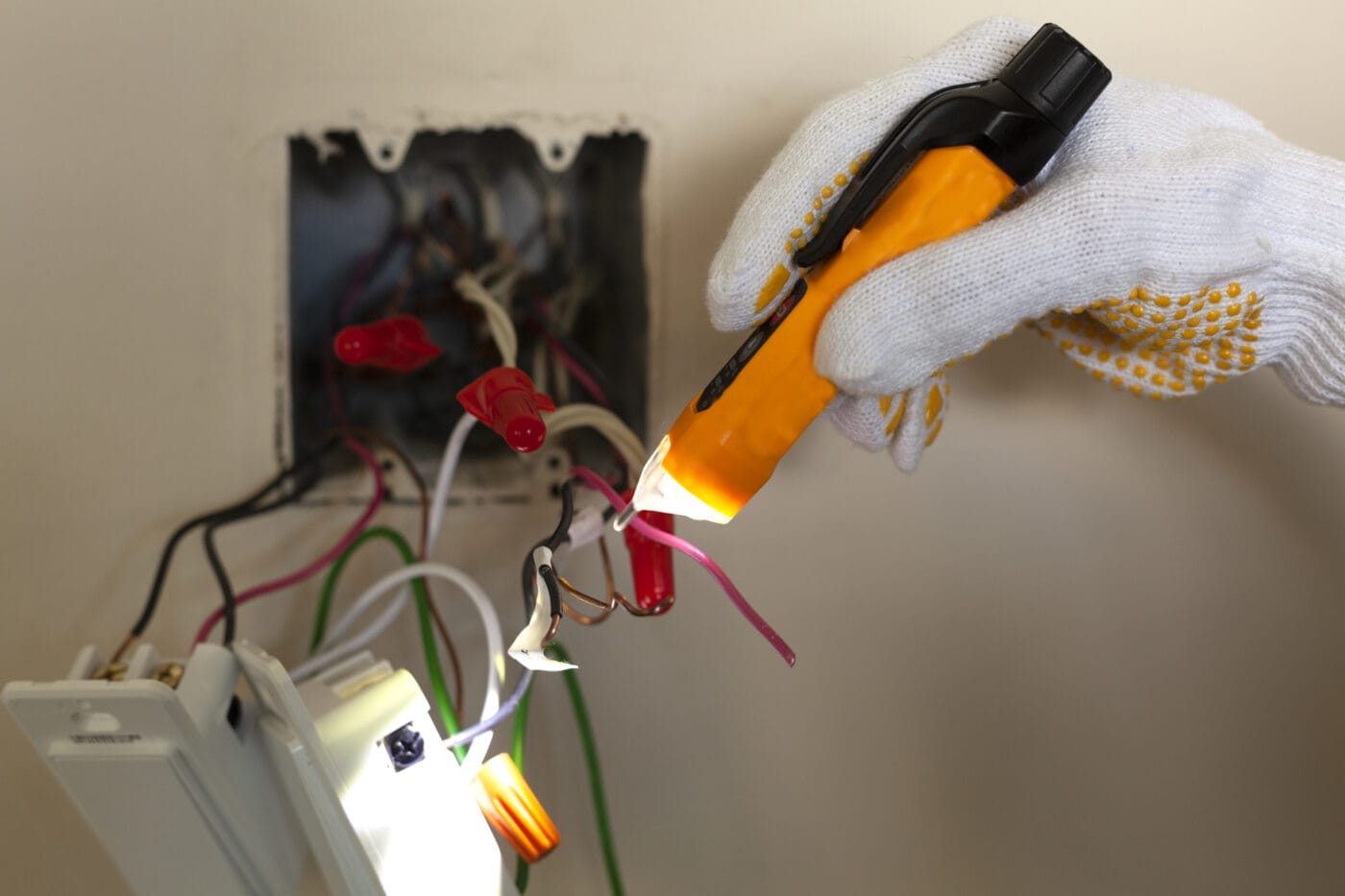 A gloved hand uses a voltage tester to check wiring inside an electrical box.
