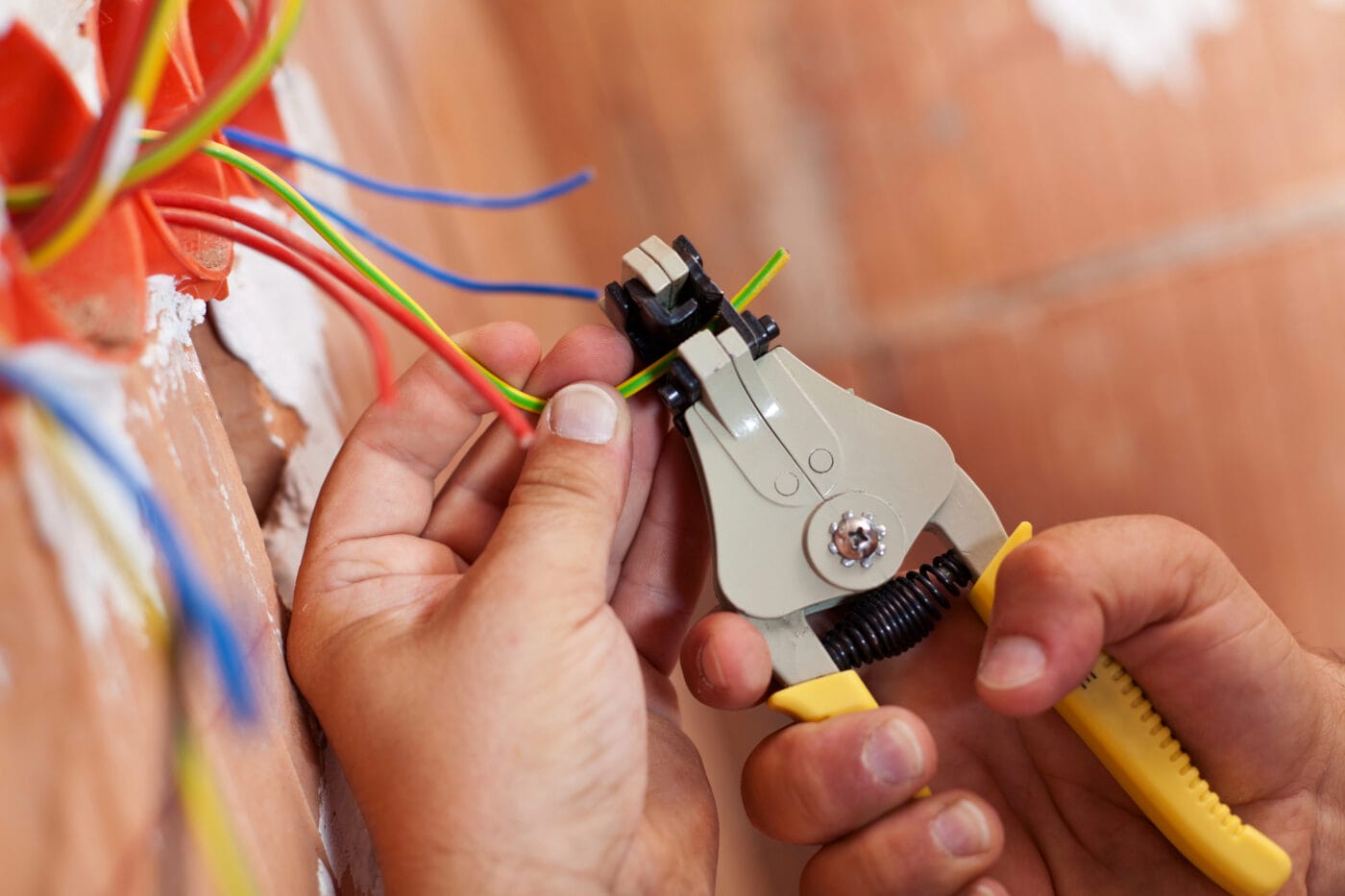 Hands using a wire stripping tool on electrical wires in front of a wall socket.