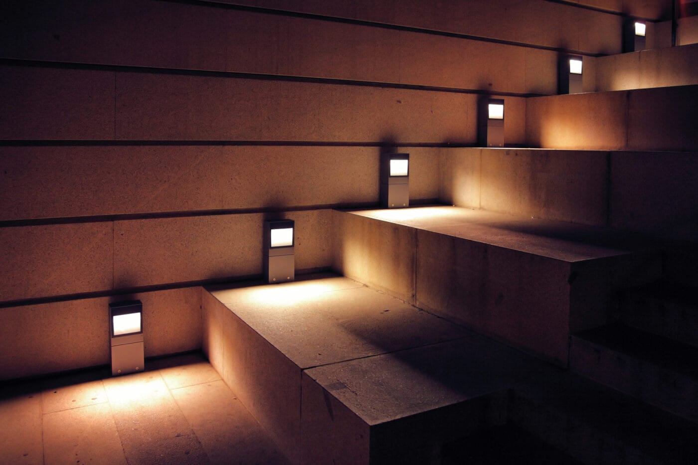 Dimly lit stone steps with rectangular wall lamps, casting soft glows and shadows on the horizontal and vertical surfaces.